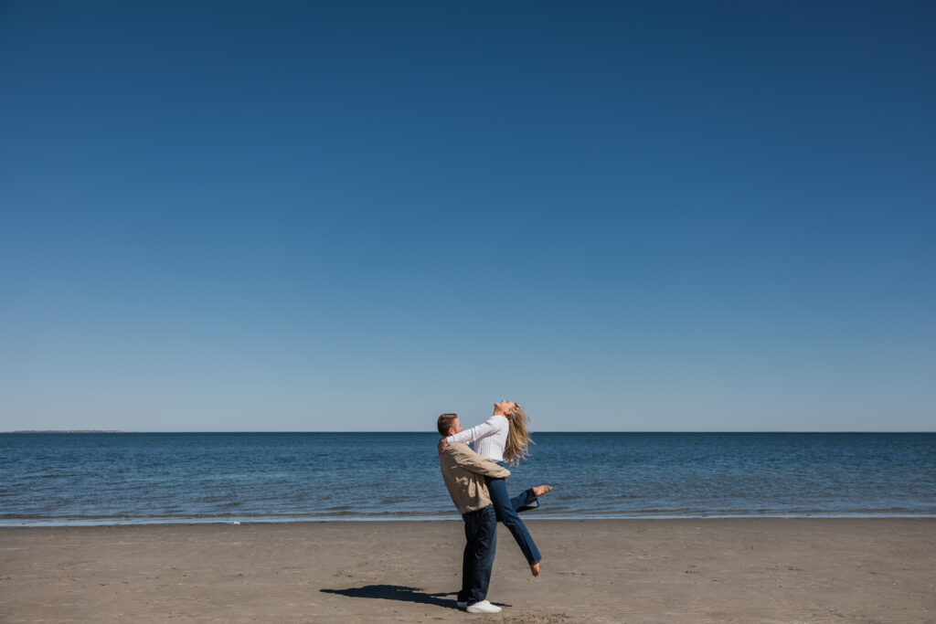 two people one picking someone up with the beach and blue sky behind them